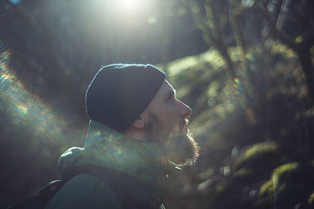 Bearded man in outdoor gear and beanie gazes thoughtfully upwards in a dimly lit forest setting, with sunbeams filtering through bare winter trees.