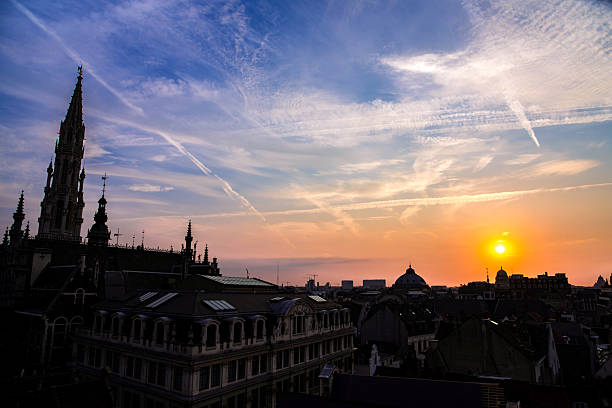 A stunning sunset over Brussels, Belgium, with the skyline silhouetted against a colorful evening sky. The Brussels Town Hall tower, an iconic Gothic structure located in Grand Place, stands prominently on the left.