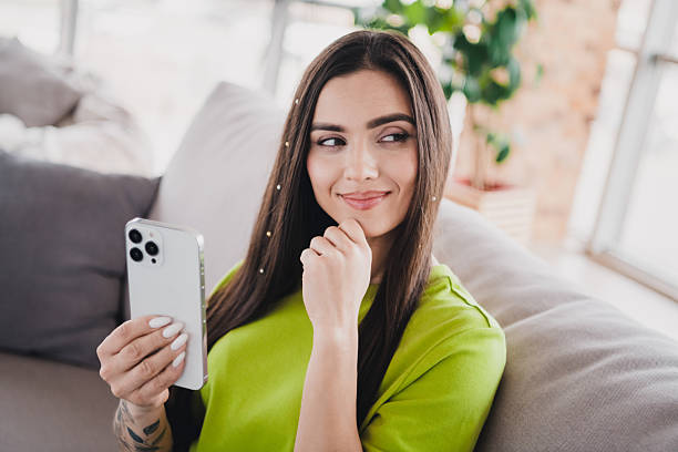 istock Young woman sitting comfortably on a sofa indoors, smiling while holding a smartphone, dressed in a green shirt 2208079412
