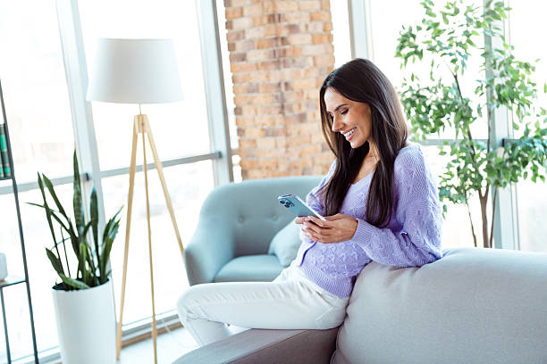 istock Young woman using a smartphone while relaxing on a cozy sofa in a modern living room with daytime sunlight and stylish decor 2208082538