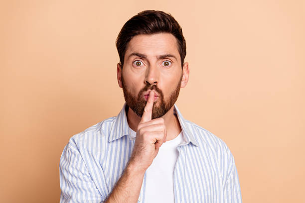 istock Young man with beard making a quiet hand gesture against a beige background, wearing a striped shirt, showcasing casual style 2208083425