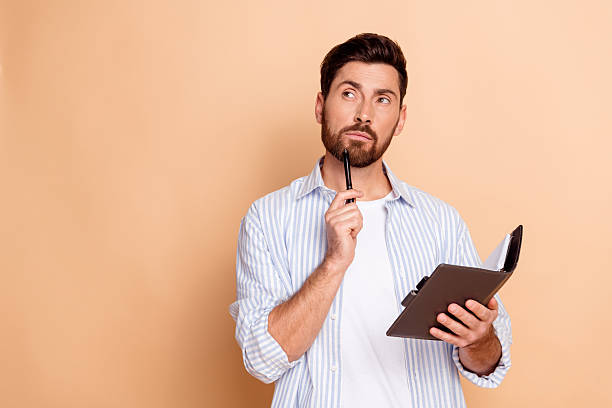 istock Thoughtful young man with a beard holding a notebook and pen in hand against a beige background 2208083449