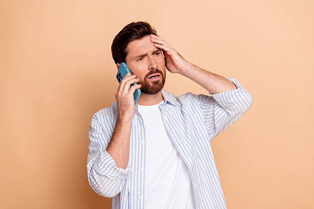 istock Young bearded man in striped shirt making a concerned gesture while talking on phone against beige background for lifestyle concept 2208083466