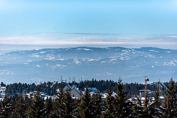 A captivating winter scene featuring a mountain range under a clear sky, with forested slopes and some housing in the foreground. The image showcases the serenity and majesty of nature during winter. A mixture of modern and natural elements adds depth to the overall composition, creating a sense of peaceful coexistence between development and the wilderness.