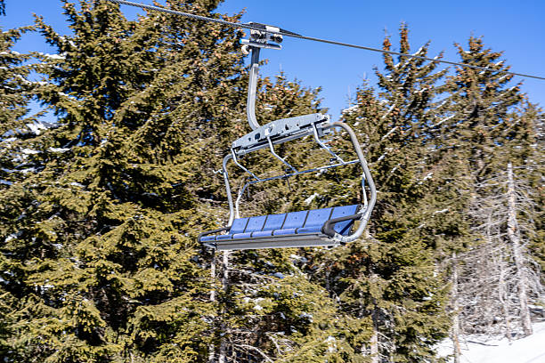 This image features a ski lift chair hanging in a tranquil winter mountain environment. The foreground focuses on the modern ski lift, while the background highlights lush evergreen trees, lightly dusted with snow, under a vivid, clear blue sky. This scene evokes adventure, exploration, and a peaceful connection with nature.