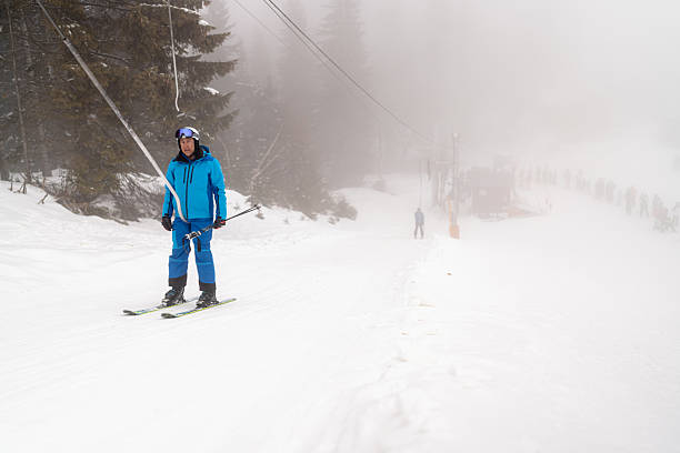 A skier in blue sportswear uses a tow lift to ascend a snow-covered slope. The scene is surrounded by tall trees and obscured by mist, creating a wintery atmosphere. In the distance, other skiers stand in line, waiting for their turn. The image conveys a serene winter sports experience.