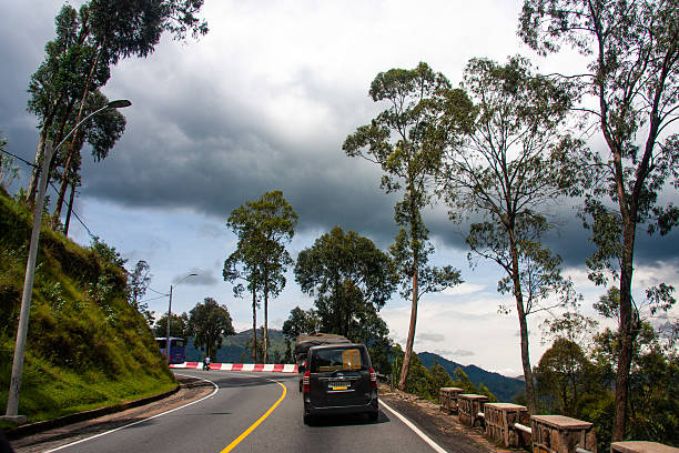 Editorial photograph of a road winding through a mountainous landscape in Rwanda. The image features a paved road with a yellow center line, curving through hills covered with trees and vegetation. A dark-colored vehicle is traveling along the road. The sky is overcast with dramatic dark clouds, creating a dynamic atmosphere. The road has a guardrail along the edge, and the landscape includes trees and visible mountain slopes. The photograph captures a moment of travel and the natural beauty of a mountain region in Rwanda.