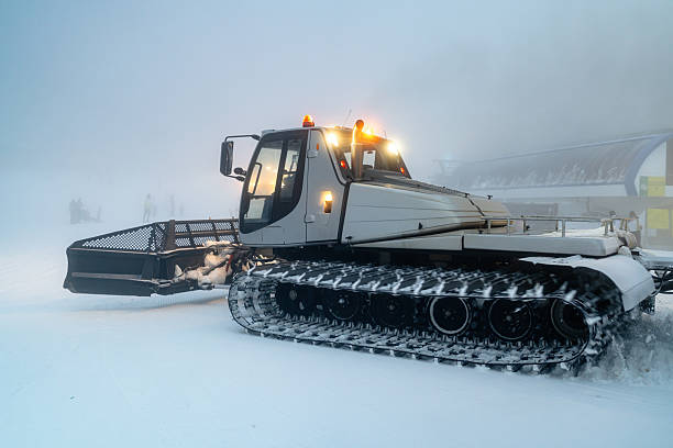 A snowcat vehicle grooming a ski slope in a snowy and foggy outdoor setting. The machine is actively preparing the surface for skiing under low visibility conditions, emphasizing its role in winter sports infrastructure.