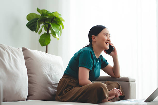 Asian woman enjoyed talking on phone while working on laptop at home. Happy woman working from home.