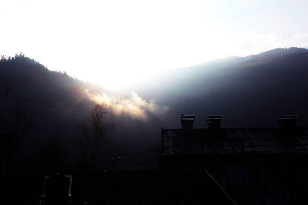 The sun shines above a layer of fog that partially covers the mountains, with houses visible in the foreground