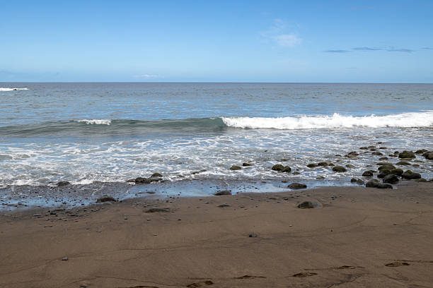 Beach with sand and pebbles on the coast of Atlantic ocean. Small waves. Blue sky with light clouds. El Burrero, Las Palmas, Gran Canaria, Canary Islands, Spain.