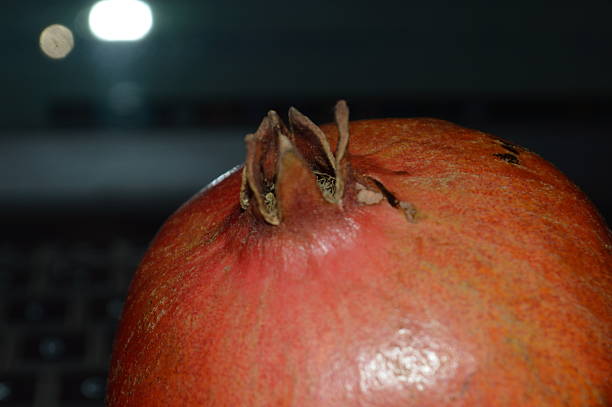 Close-up shot of a vibrant pomegranate fruit resting on a table, with dramatic dark shadows cast below. The rich colors and shadows create a striking