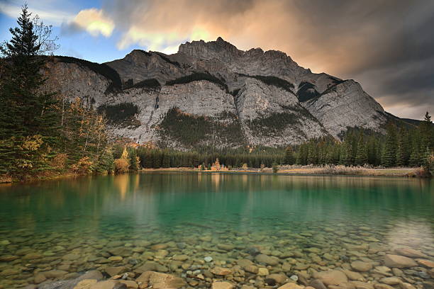 West-northwestward view of the 2998 m.high Cascade Mountain from the Cascade River and Pond over conifer forest and balsam poplars in autumn foliage under colorful cloudy sky at dusk. Banff-AB-Canada.