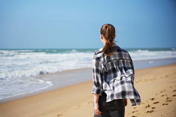 Young woman in plaid shirt and ponytail stands on sandy beach, looking at waves under clear blue sky on windy day.