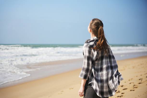 Young woman in plaid shirt with ponytail stands near ocean, looking up and soaking in a calm, breezy beach moment.