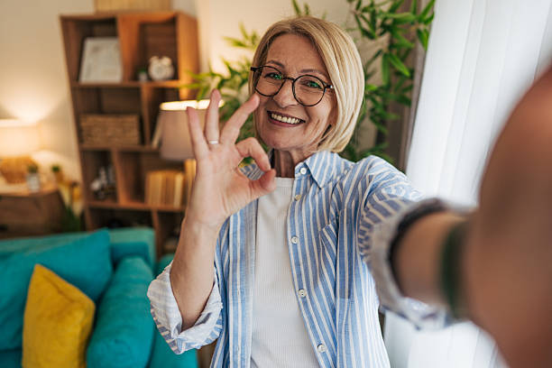 istock Smiling senior woman making ok sign while taking selfie at home 2208100742