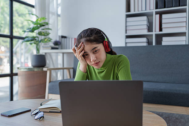 Woman sitting at a table with a laptop and headphones engaged in e-learning while maintaining a serious demeanor at home