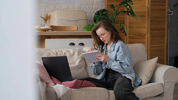 istock A woman in a denim jacket works on her laptop and takes notes while sitting on a comfortable couch in her cozy living room. This image emphasizes the flexibility and comfort of remote work in a home environment. 2208101429
