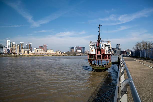 A rusting, abandoned boat sits partially submerged along the River Thames, with modern London apartments towering in the background under a clear blue sky.