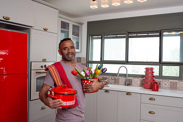 A cheerful man poses in a cozy home kitchen, holding a red cooking pot in one hand and a polka dot container filled with colorful tulips in the other. Dressed casually with a towel over his shoulder, he stands in front of a sunny window and patterned tile backsplash, adding a playful, seasonal touch to the kitchen setting.