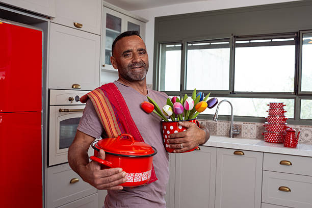 A smiling man poses in a cheerful kitchen, holding a red cooking pot and a red polka dot container filled with colorful tulips. Dressed casually with a dish towel over his shoulder, he stands in front of a bright window and patterned tile backsplash, bringing a sense of freshness, warmth, and springtime energy to the home environment.