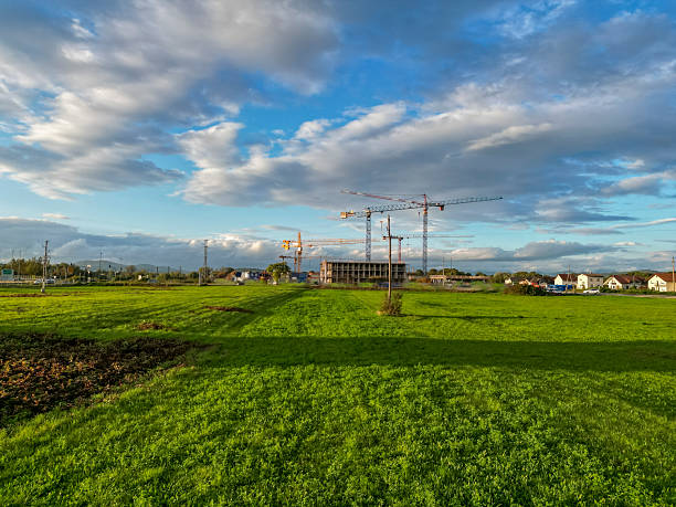 A sprawling green landscape with a building under construction, tall cranes, and a bright blue sky with scattered white clouds at daytime.