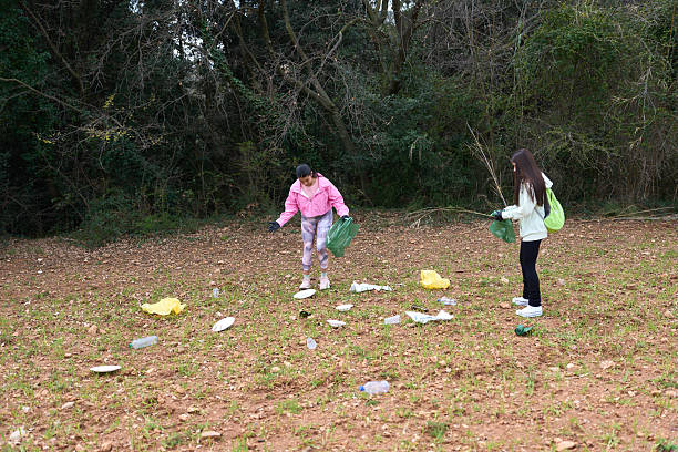 A family of three wearing gloves cleans a grassy park area by picking up trash in black bags during daylight. They are focused on maintaining the outdoor environment in a natural setting with trees and water nearby.