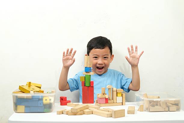A cheerful young boy in a blue shirt is playing with wooden building blocks, constructing a structure. He is focused and engaged, showcasing creativity and learning through play in a bright indoor setting.