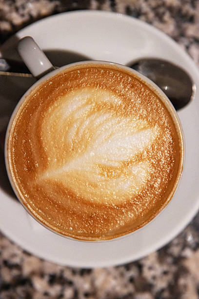 Top view of a creamy cappuccino with elegant latte art in a white ceramic cup with a spoon on a saucer, on a granite table. Coffee culture, creativity, barista skills concept and cozy cafe atmosphere