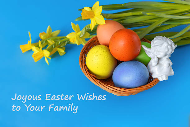 Colorful Easter eggs in wicker basket decorated with ceramic angel figure and fresh yellow daffodils on blue background. Card with wishes.