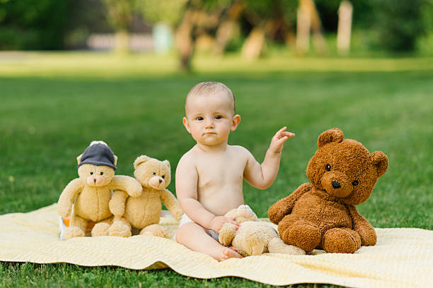 A baby sitting on a blanket outdoors, smiling and playing with teddy bears, enjoying a lively moment surrounded by nature and greenery.