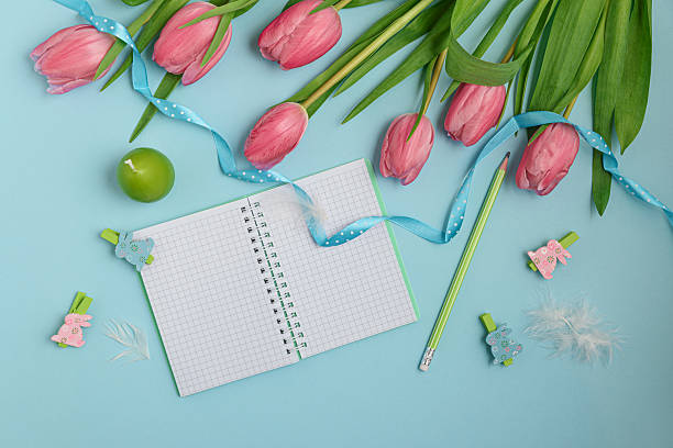 A pastel Easter scene featuring a bouquet of pink tulips adorned with a delicate blue ribbon, a notebook, wooden rabbit figurines, and soft feathers. Perfect for celebrating the Easter holiday.