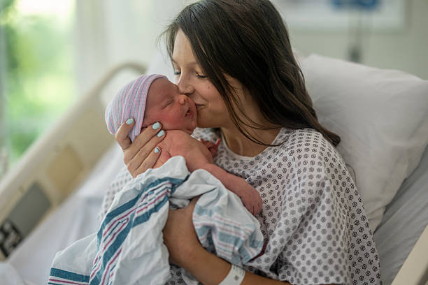 A new Mother sits up in her hospital bed shortly after delivery as she hols her baby close and kisses his cheek.  She is wearing a hospital gown and has a gentle smile on her face as she spends time bonding with her baby.
