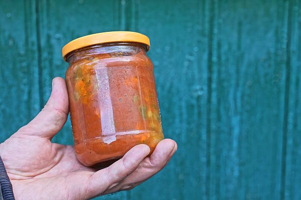 hand holds one closed glass jar with red lecho from canned pepper against the background of a green wall