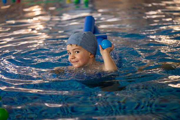 Young boy learning to swim with a pool noodle and playing with a blue ball in an indoor swimming pool
