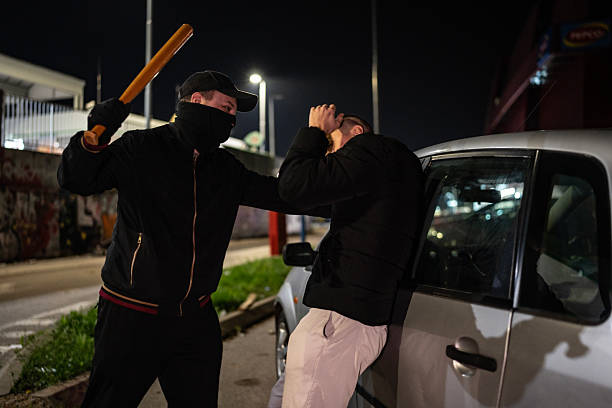 A man wearing a mask and holding a bat confronts another person outdoors near a vehicle at night, suggesting themes of danger, confrontation, and urban tension.