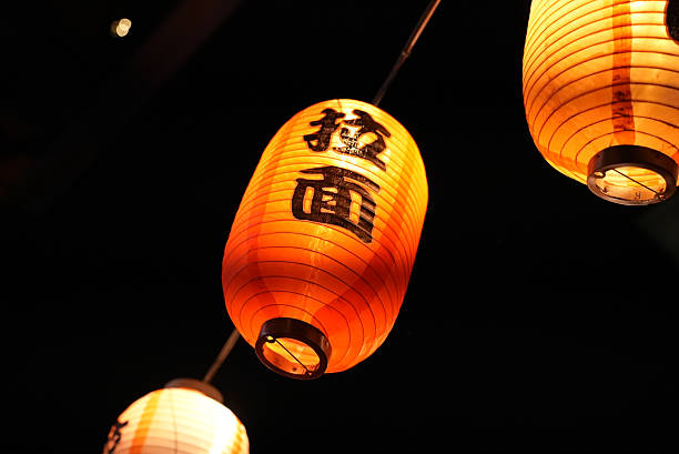 A colorful shot of lanterns hanging outside a traditional Japanese ramen shop, glowing softly in the evening. The lanterns, emblazoned with Japanese characters, create a warm, inviting atmosphere, adding to the charm of the street food culture in Japan.