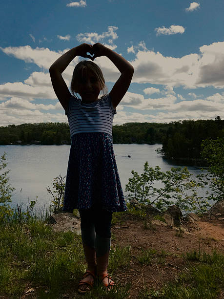 A young girl, around 6-7 years old, stands high on a bluff overlooking a peaceful lake. With her arms raised, she forms a heart shape with her hands, silhouetted against a moody yet beautiful sky. The scene captures a sense of wonder, freedom, and connection to nature. The light is soft and atmospheric, creating a quiet, emotional portrait in a dramatic natural setting. Photo taken at Morgan Park Campground in Northern Wisconsin.