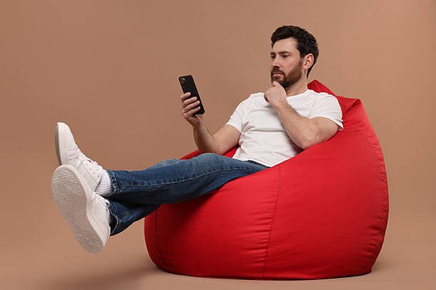 istock Handsome man with smartphone sitting on bean bag against light brown background 2208209375