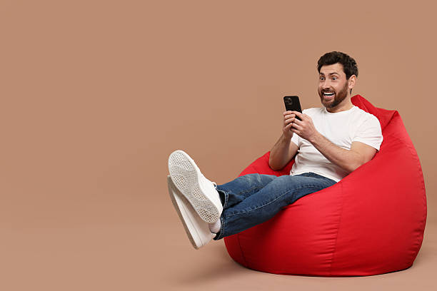 istock Happy man with smartphone sitting on bean bag against light brown background. Space for text 2208209406