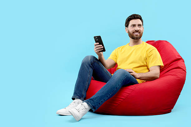 istock Smiling man with smartphone sitting on bean bag against light blue background. Space for text 2208209460