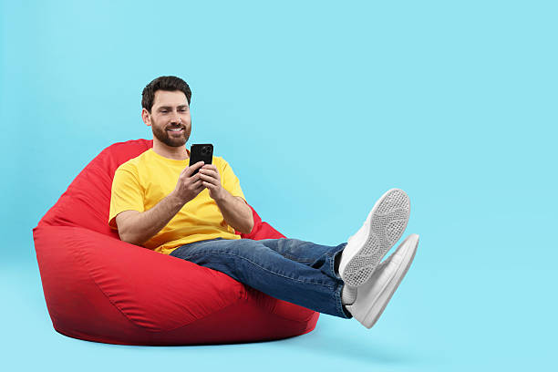 istock Smiling man with smartphone sitting on bean bag against light blue background. Space for text 2208209473