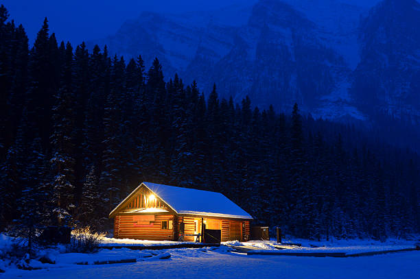 A small wooden cabin covered in snow sits peacefully in a winter landscape at night. The surrounding trees are heavily laden with snow, and gentle snowfall adds to the serene and magical atmosphere. The warm glow of the sky contrasts beautifully with the cold, white snow, creating a cozy yet isolated scene in nature.