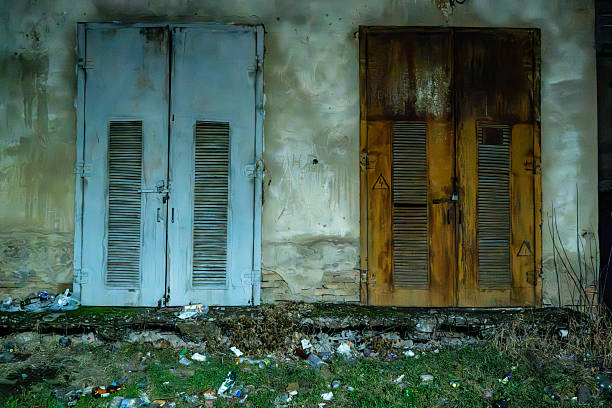 A close-up view of an old tropical-style building exterior featuring a faded red wooden window with louvered shutters. The concrete walls show signs of weathering, with patches of moss, algae, and peeling paint in shades of grey and white. The structure appears abandoned or aged, evoking a sense of rustic decay in a coastal or urban setting. No people are present. This image suits commercial uses in travel articles on historic architecture, lifestyle features on renovation projects, or editorial content about urban preservation and tropical environments. It can illustrate themes of timeworn structures in regions like the Caribbean or Southeast Asia, ideal for book covers, websites, or magazines focusing on architecture and decay.