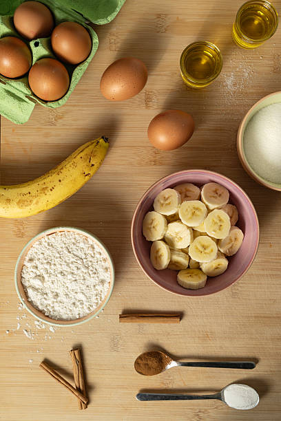 op-down photograph showcasing the essential ingredients for making a banana cake: egg, oil, banana, flour, cinnamon, yeast, and sugar. The natural wooden background and organized arrangement of the ingredients create a warm and inviting atmosphere, perfect for a homemade recipe.