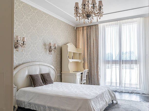 istock cozy retro-style bedroom with beige wallpaper, bed with pillows and bedspread, bedside table, lamp on table, wall lamp. bedroom interior. vintage chandelier with crystals, chest of drawers. 2208260012