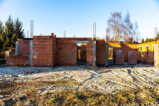 A partially built house structure made of red bricks with visible rebar and construction materials, photographed in daylight on a sandy plot in Poland