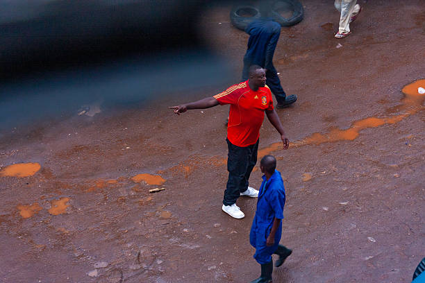 An everyday urban scene in Kigali, Rwanda, shows a man in a red sports jersey gesturing while engaging with others on a wet, unpaved street. The surrounding individuals, dressed in work attire, add to the dynamic atmosphere of the city. The image reflects the rhythm of daily life in Kigali, highlighting human interaction and movement in an urban environment.
