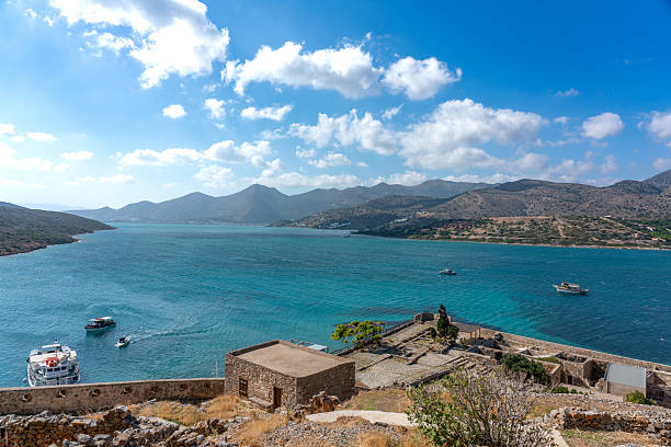 A breathtaking coastal scene showcasing clear turquoise waters, boats, and mountains under a bright sky. Ancient stone ruins in the foreground add historical charm