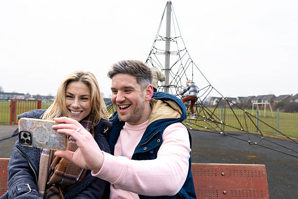 A wide shot of a mother and father sitting down on a bench at a public park in Darlington. The man is holding a mobile phone to take a selfie of him and his wife while smiling. Their children are playing on a climbing frame behind them in the park.\n\nVideos are available similar to this scenario.